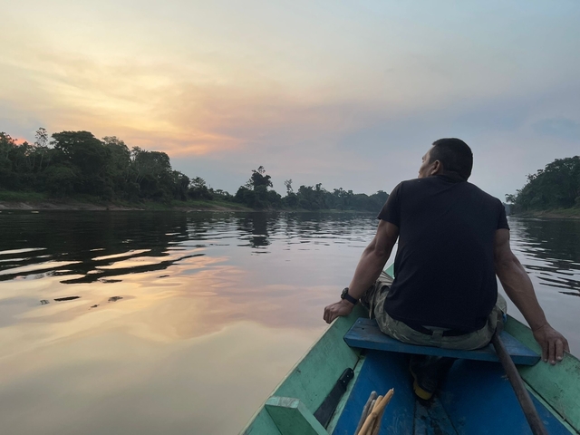 A person sitting on a boat looking at a sunset over a river with trees on the shore.