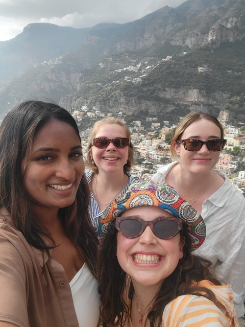 Four women smiling with a scenic town and cliffs in the background.