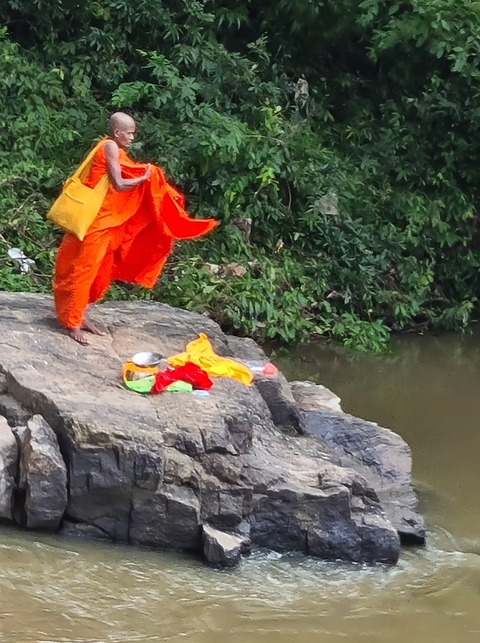 Monk in orange robes near a river, carrying vibrant textiles.