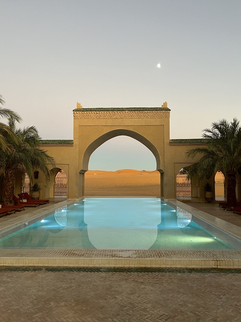A pool leading to a desert landscape with an arched structure and palm trees.