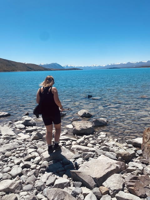 Person standing by the edge of a clear blue lake with mountains in the background.
