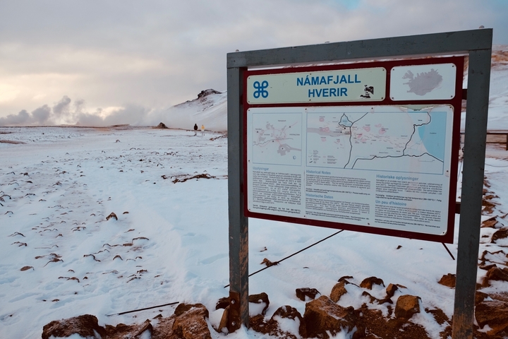 Informational sign in a snowy landscape with geothermal steam in the distance.