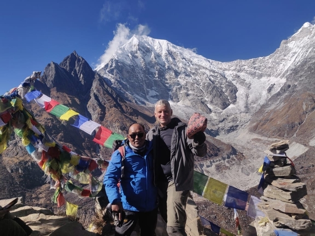 Two people posing in the mountains with prayer flags and snow-covered peaks.
