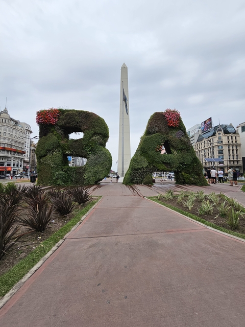 Topiary letters BA with a tall obelisk and city buildings in the background.