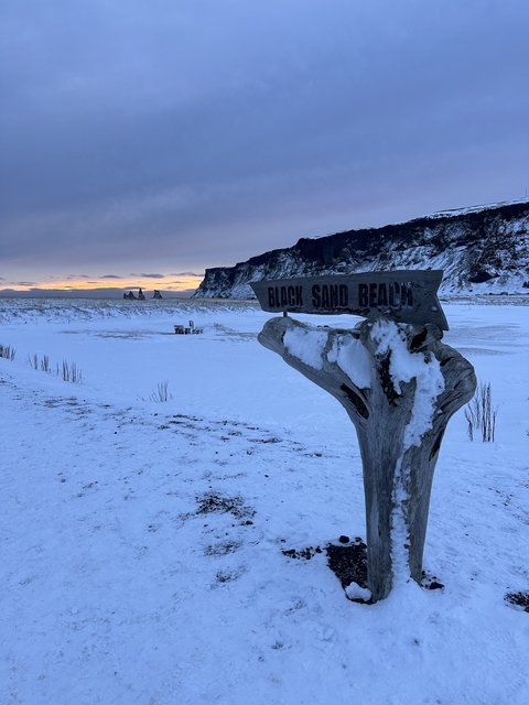 A snowy landscape with a sign pointing to Black Sand Beach.
