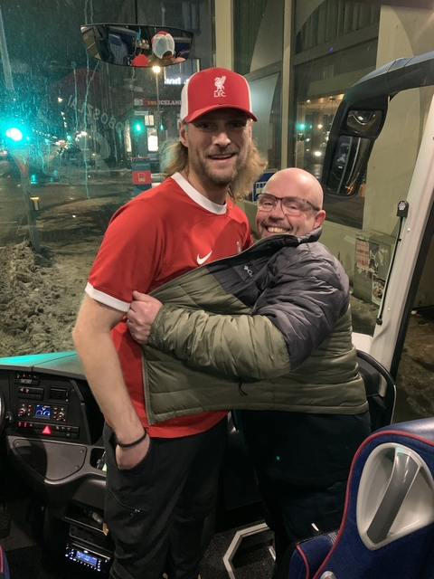 Two men posing together inside a vehicle, one wearing a red shirt.