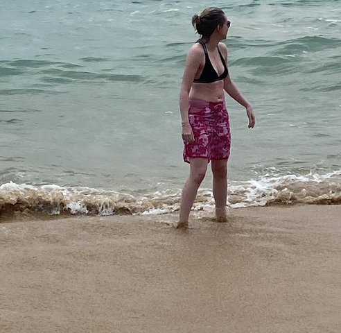 Partial view of a person standing on a sandy beach with waves touching their feet.
