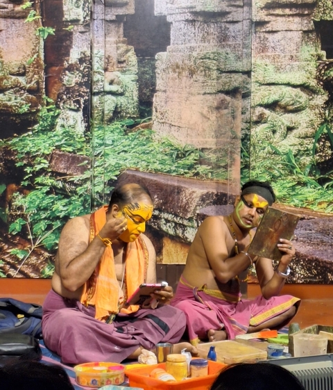 Two artists applying face paint, sitting in front of a decorative background.