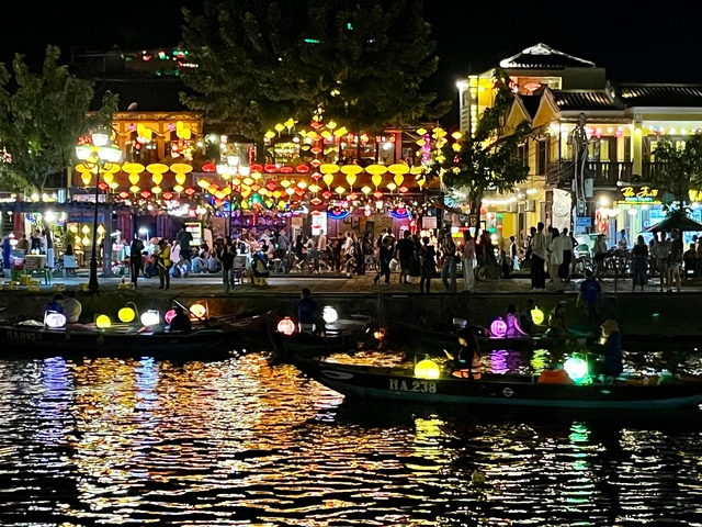Night view of a bustling street with illuminated lanterns and boats on the water.