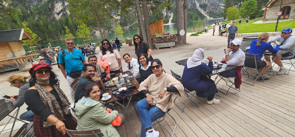 Large travel group enjoys drinks at outdoor lakeside café with alpine lake and forested mountains in the background.