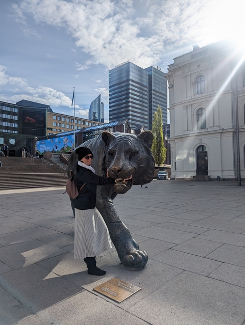 Person posing with a large tiger sculpture in a city square.