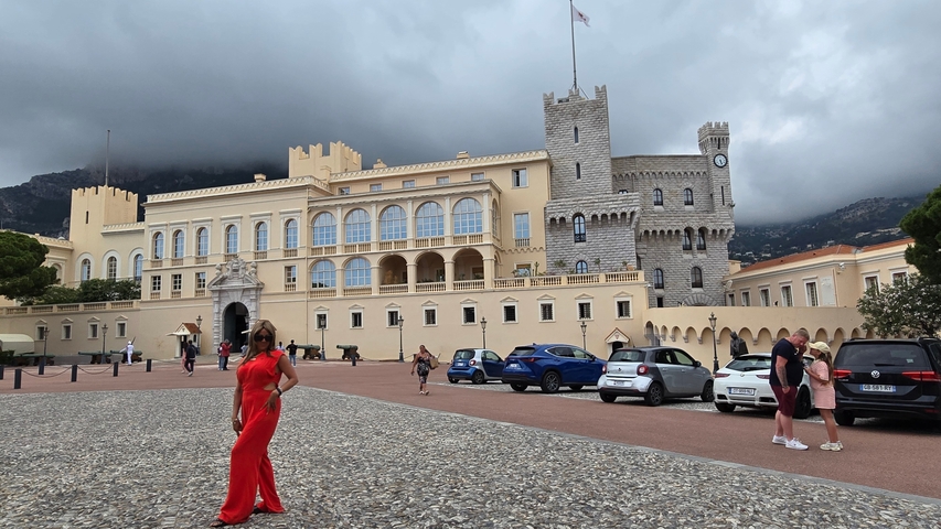 A woman posing in front of a historic palace with parked cars and cloudy sky.