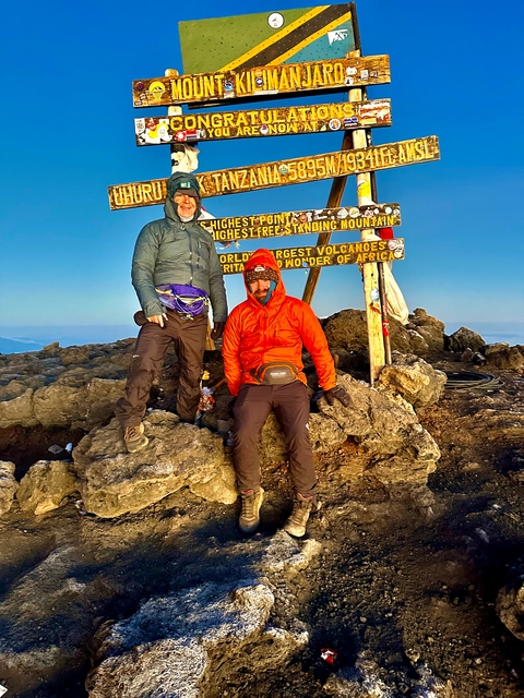 Two individuals at a mountain peak summit with signs.