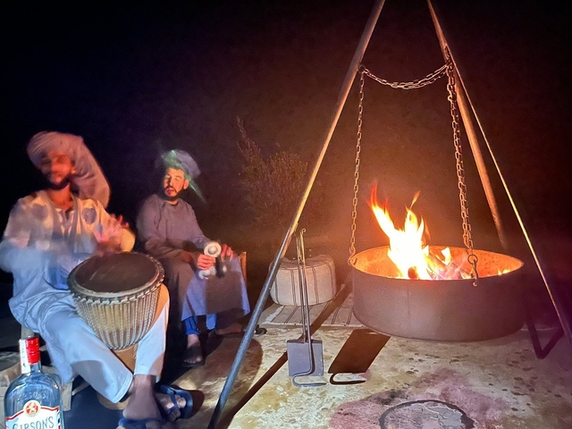 Two people drumming and sitting beside a fire under the night sky.