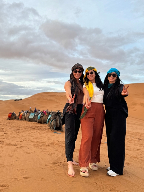 Three women posing with peace signs in front of camels in the desert.