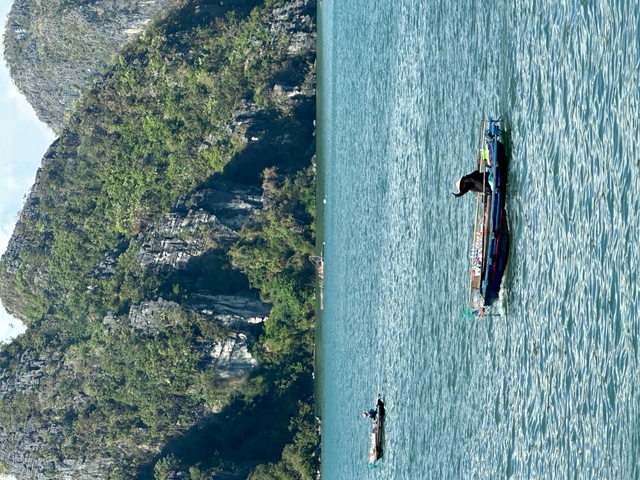 Long wooden boat gliding across turquoise water with towering limestone cliffs covered in greenery behind it.
