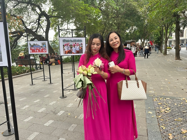 Two smiling women in bright pink ao dai holding lotus flowers on a tree-lined sidewalk gallery.