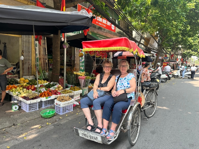 Two tourists seated in a colorful cycle-rickshaw on a busy Vietnamese market street lined with fruit stalls.