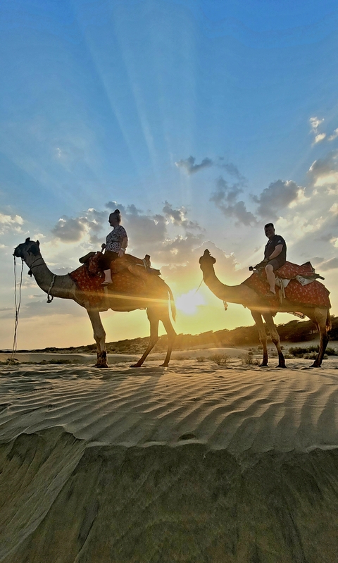 Two people riding camels in the desert at sunset.