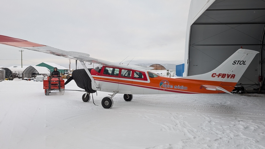 Small propeller aircraft with skis parked on snowy airfield while staff load equipment.