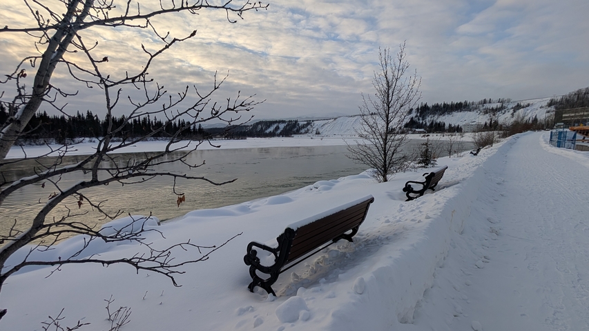 Snow-covered riverside path with empty benches overlooking a partially frozen river under textured clouds.