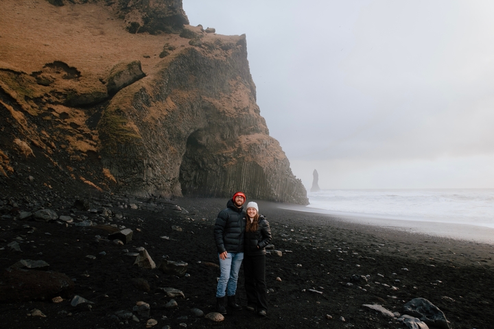 Two people standing on a black sand beach with rocky cliffs and a distant sea stack.