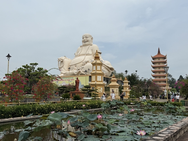 Large statue of Buddha with a temple pagoda, surrounded by people and lotus flowers.