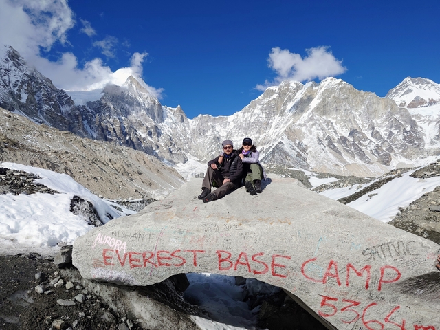 Two people sitting on a rock labeled 'Everest Base Camp' with snowy peaks.