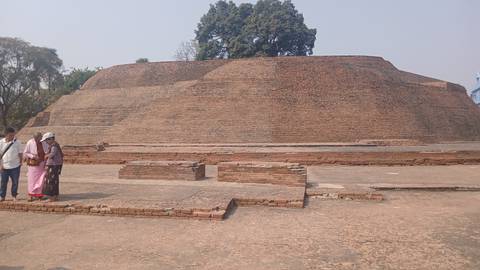 People standing near a large brick stupa with steps, surrounded by trees.