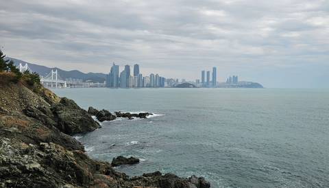 Seaside cityscape with tall buildings and a bridge under a cloudy sky.