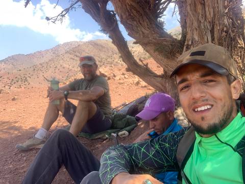 Three hikers resting under a tree in a mountainous area.