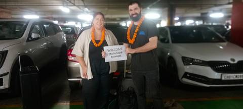 Smiling couple with garlands holding a welcome sign in a parking lot.
