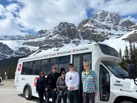 Group of people posing in front of a tour bus with snow-capped mountains in the background.