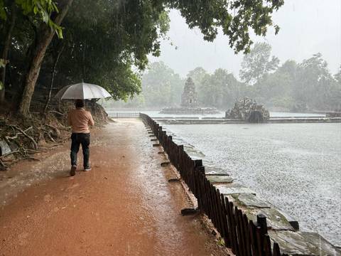 Person walking with umbrella near ancient ruins by a lake during rain.