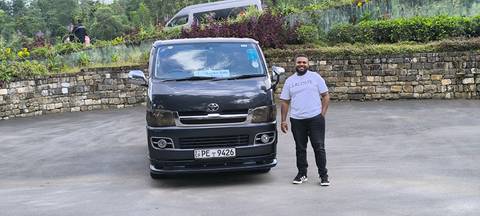 Man posing beside a van in a lush green setting.