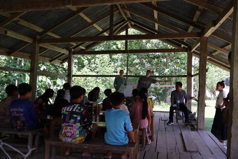 A group of people attending an outdoor class or meeting under a wooden shelter.