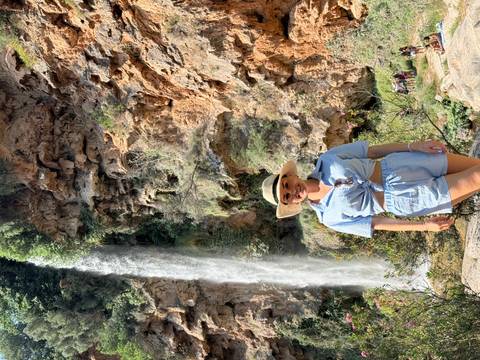Woman posing in front of a waterfall within a rocky landscape.
