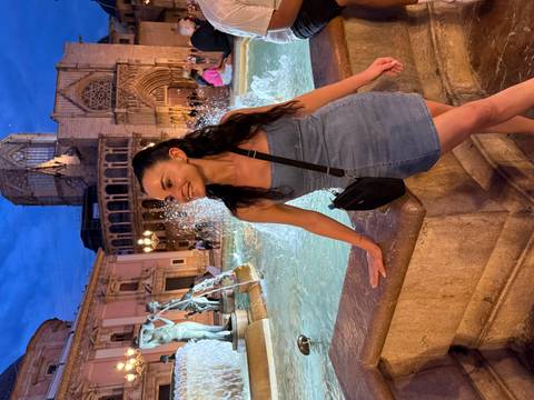 Woman posing by a fountain at a lively night setting in a historic area.