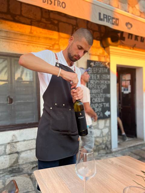 Person opening a bottle of wine with a restaurant chalkboard menu in the background.