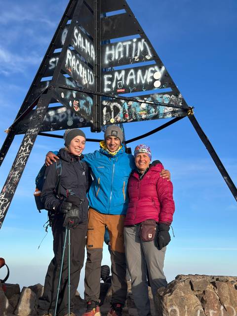 Three people posing under the sign at a mountain peak.