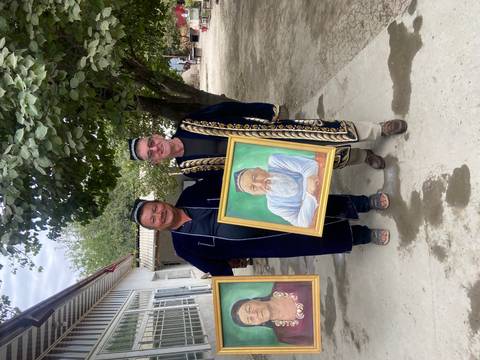 Two men holding portraits, standing outdoors under a tree.