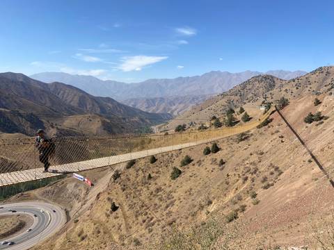 A person crossing a long suspension bridge over a deep valley with mountains in the background.