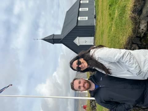 Two people in front of a black church with grass and wooden structure.