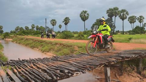 Motorcyclist crossing a wooden bridge in a rural area with palm trees.