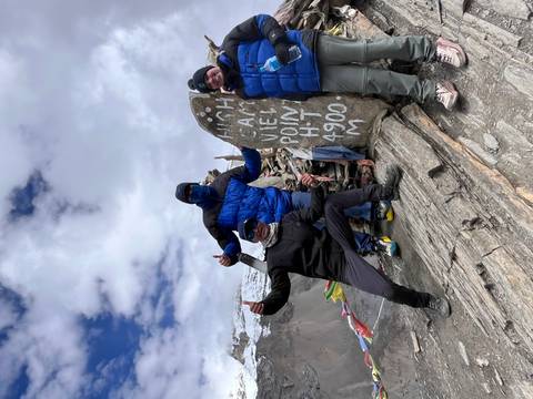 Three people posing with a stone marker saying 'High camp view point' at high altitude with mountains in the background.
