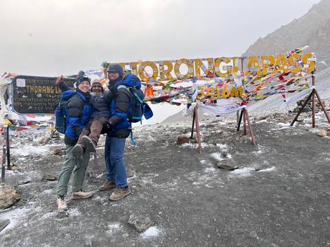 Three people with backpacks at a mountain pass with prayer flags and signs.