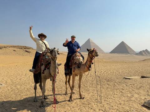 Two people on camels in front of pyramids in a desert.