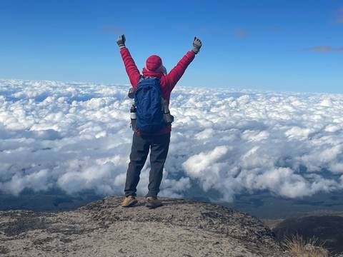 Person in red jacket standing on a mountain with arms raised above clouds.