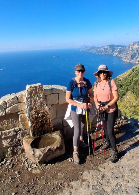 Two hikers posing with trekking poles by a stone wall.