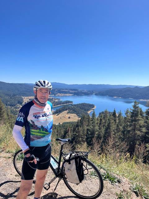 Man posing with bicycle against a scenic backdrop of a lake and mountains.